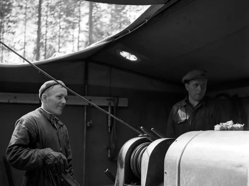 A driller guides a core barrel out of a diamond drill under a canvas shelter in a forest, 1956