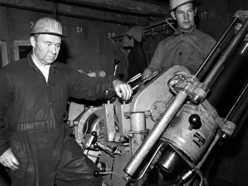 Two drillers in hard hats beside a diamond drilling machine inside a drill shed, 1962