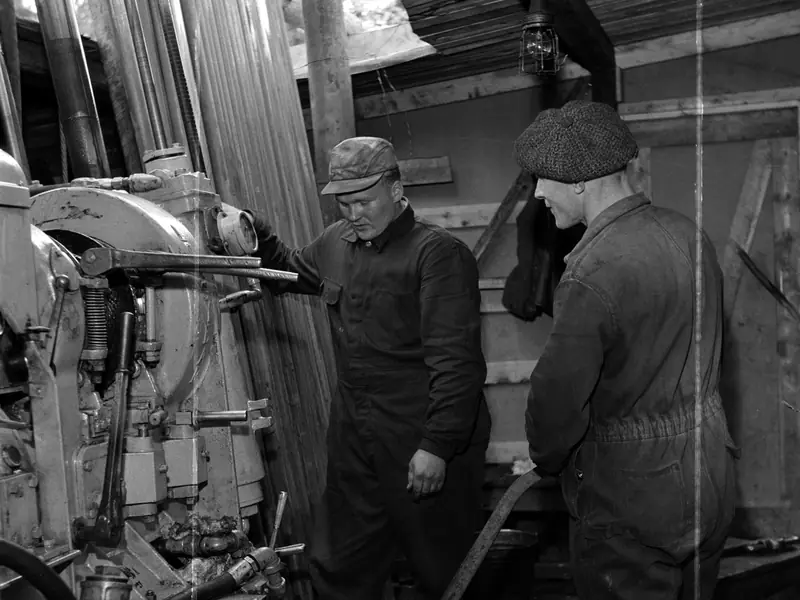Two drillers in overalls next to a diamond drill rig and stacked drill rods inside a wooden drill shed, 1962
