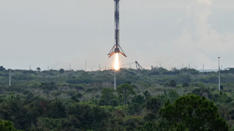 A Falcon 9 first stage descending on its engine flame toward a landing pad beyond the trees