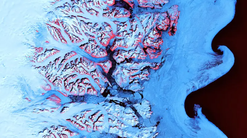 Satellite view of swirls of sea ice and icebergs off Greenland's south-east coast, with red-tinted mountain rock