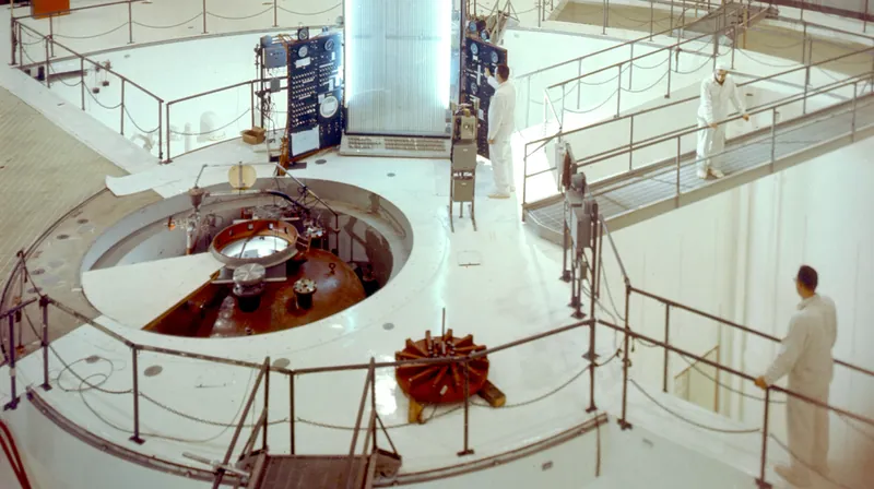 Inside a round research-reactor hall: a central pool with railings, a tall containment wall and technicians in white