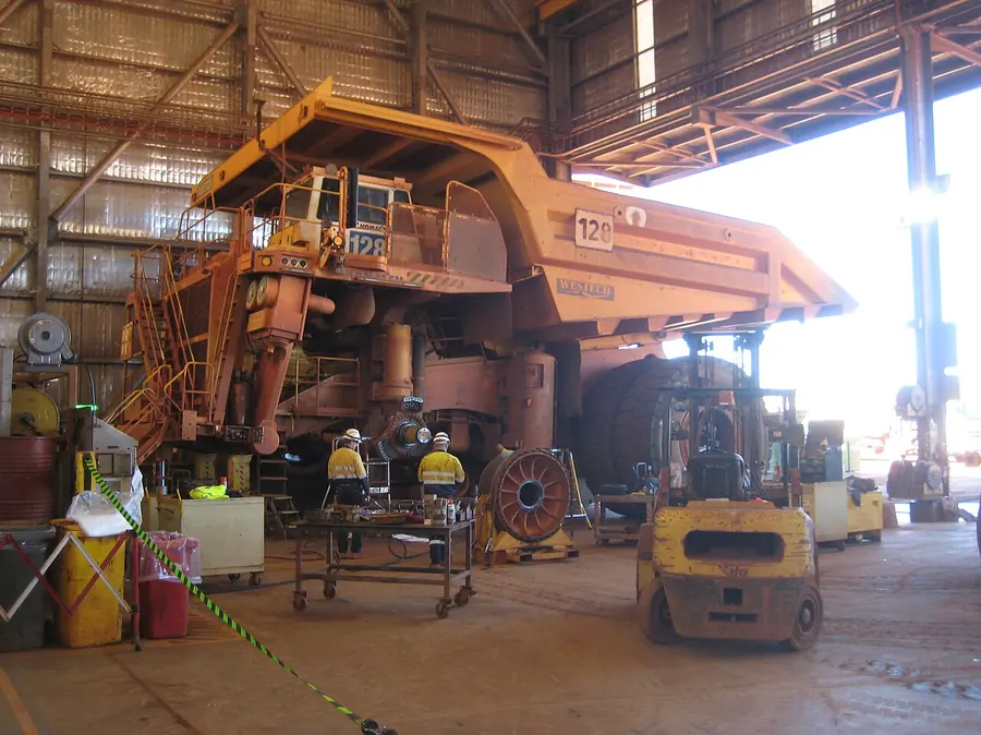 A giant haul truck with its tray raised, partly dismantled in a workshop with fitters in hi-vis working on it