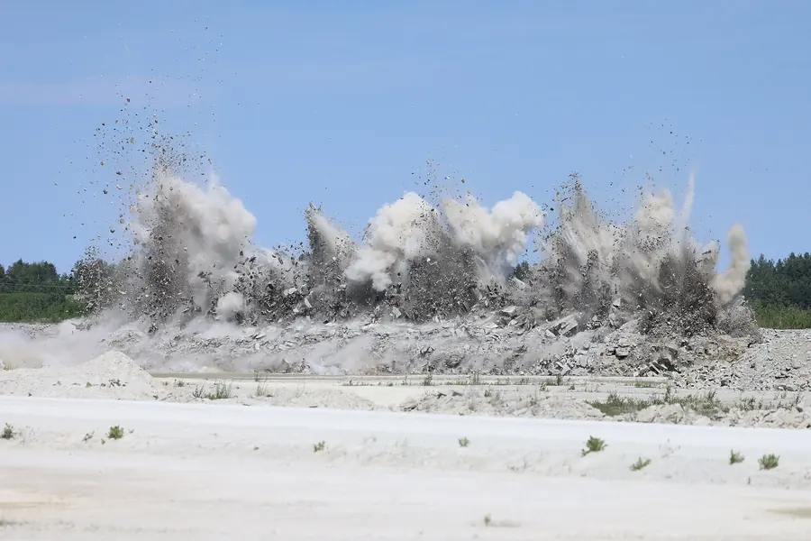 A row of rock blasts erupting along a quarry face, throwing dust and stones into the air