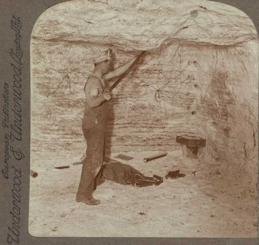 Old sepia stereograph of a miner reaching up to load a drilled hole in the wall of a salt mine