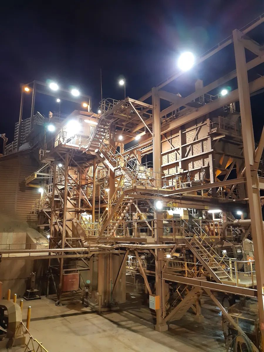 The lit steel tower of a primary crusher at night, with stairs, chutes and walkways