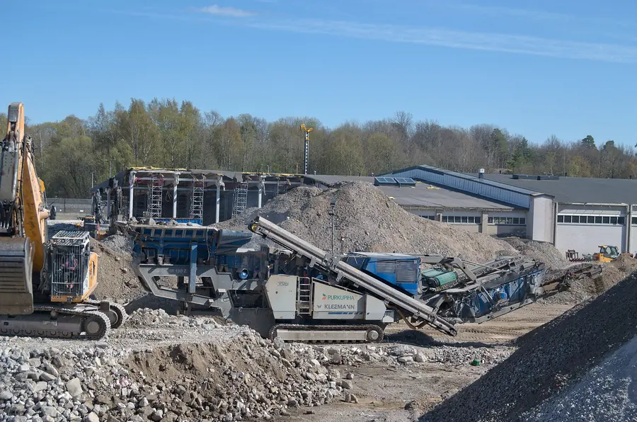 A tracked mobile crushing plant with conveyors and a stockpile, fed by an excavator
