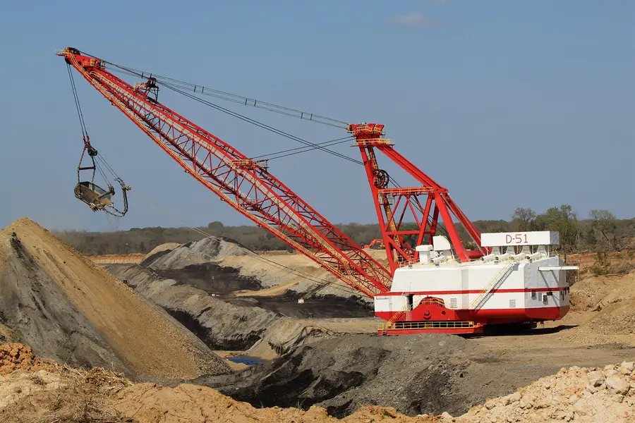 A red and white walking dragline with its bucket raised on ropes from a long lattice boom, over a lignite strip cut
