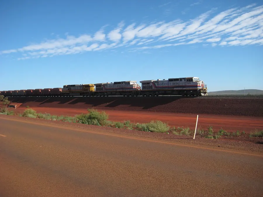 Three locomotives head a train of ore wagons across red Pilbara ground under a blue sky