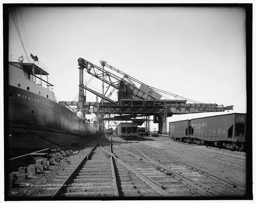 Tall steel Hulett unloaders lean over the hold of a lake freighter beside railway tracks