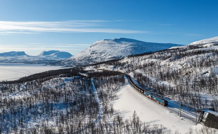 An ore train curves through snowy birch forest below white mountains