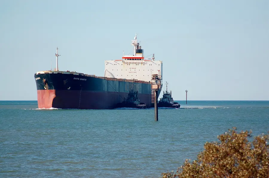 A large bulk carrier with a red hull, led by tugs, enters a harbour
