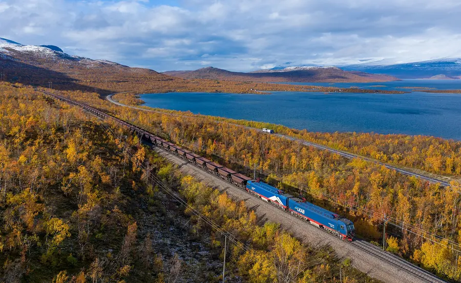 Two blue electric locomotives pull a long ore train along a lake shore through autumn birch woods