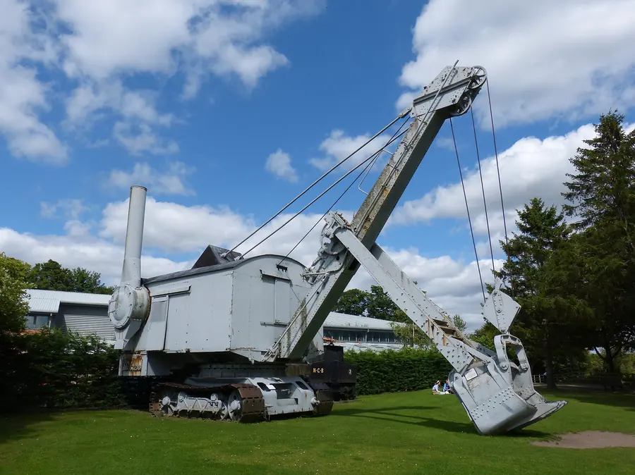 A grey preserved steam shovel with a long boom and dipper bucket on a lawn