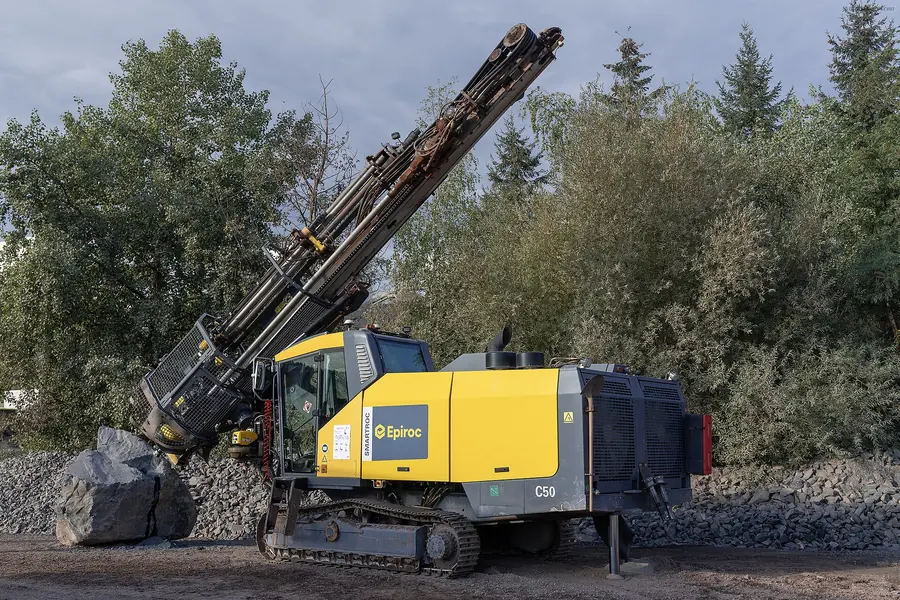 A yellow and grey tracked surface drill rig with its mast raised beside broken rock