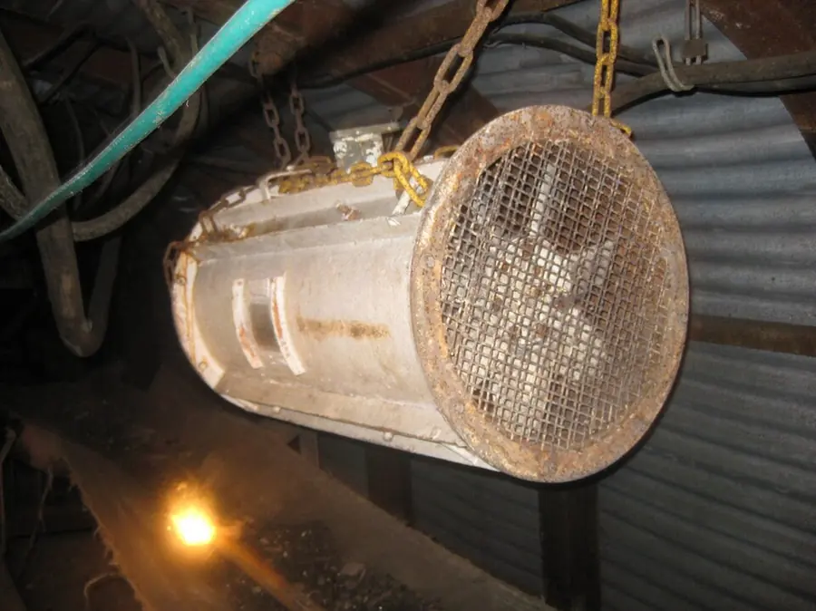 A cylindrical ventilation fan with a mesh guard hanging on chains in a mine