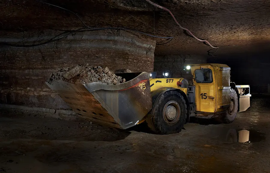 An underground loader carrying a full bucket of rock through a dark mine room
