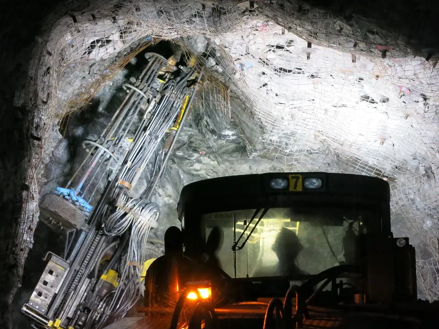 A drill rig with its boom raised in a white-walled tunnel, lights in the cab
