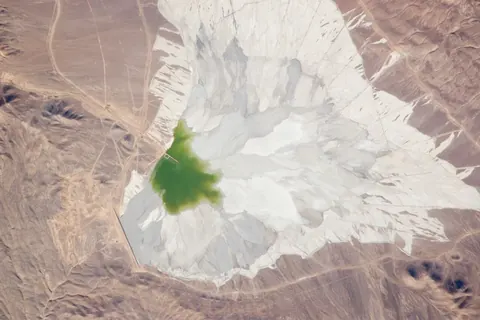 View from orbit of a pale tailings dam with a green pond in brown desert