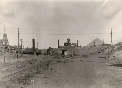 Black and white view of a dusty track leading to mine headframes, chimneys and a conical waste heap