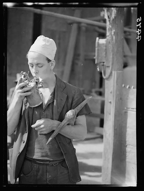 Black and white photograph of a young worker in a white cap holding a small metal piece to his mouth