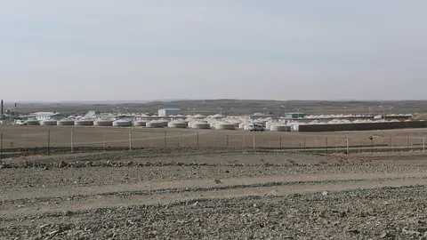 Rows of white buildings and round tents behind a fence on a flat stony plain